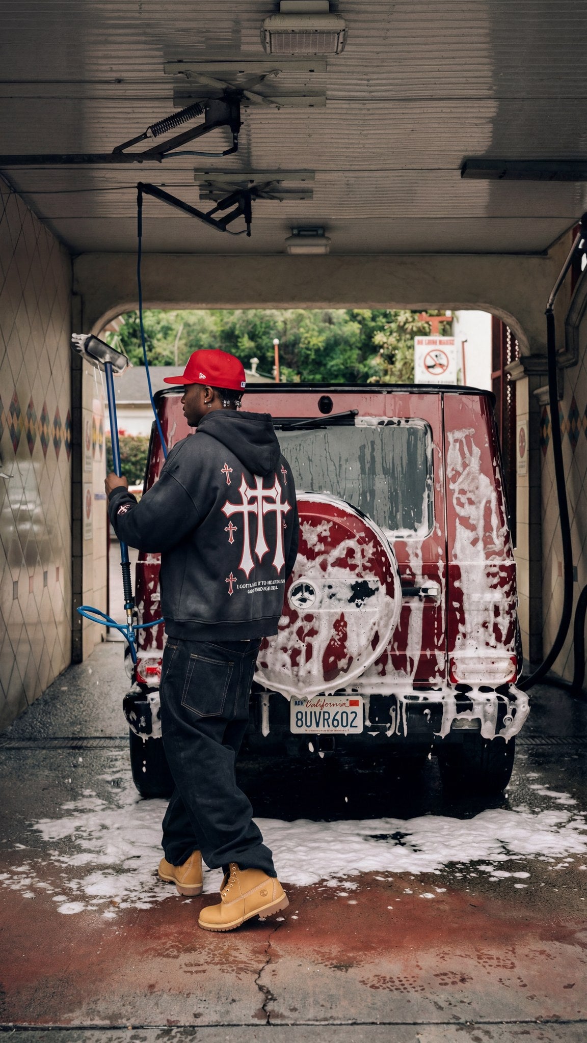 Man wearing the Saint Villains Redemption black zip hoodie showing the red and white back crosses graphic while washing a G-Wagon
