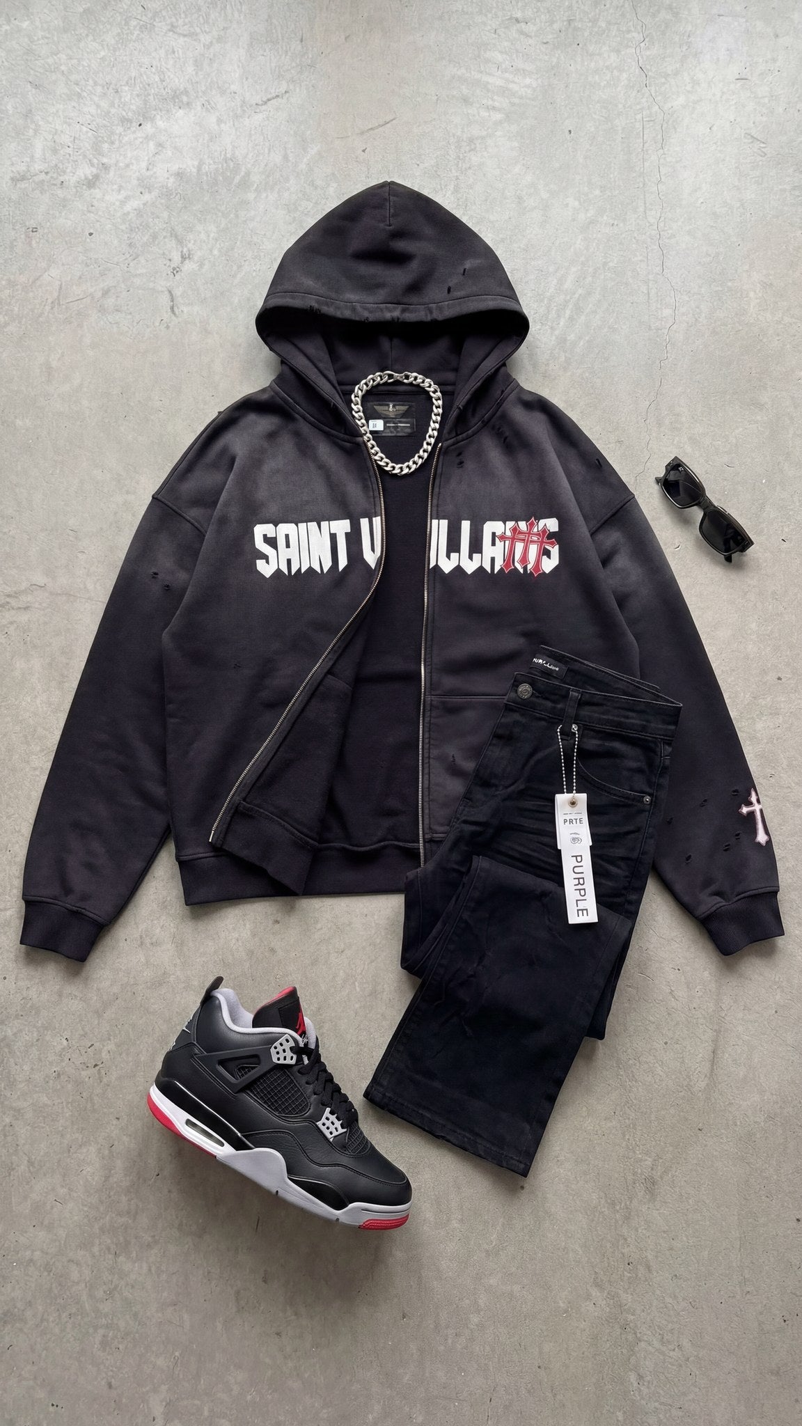 Saint Villains Redemption black zip hoodie styled flat with black denim, Jordan 4 sneakers, a silver chain and sunglasses