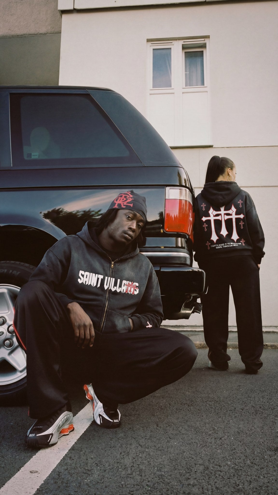 Man and woman wearing the Saint Villains Redemption black zip hoodie, showing the front lettering and the red and white back crosses graphic