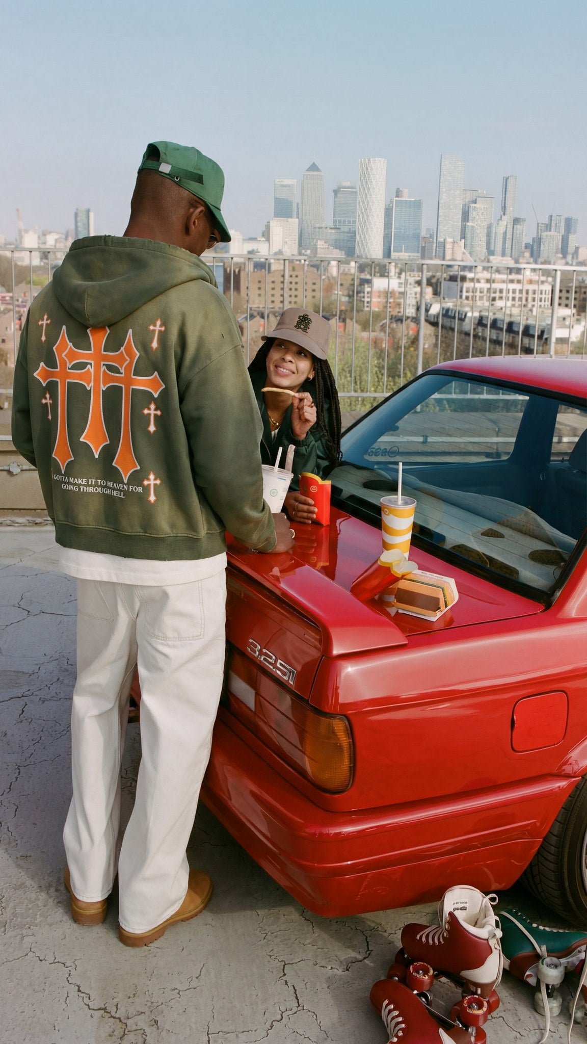 Man wearing the Saint Villains Redemption olive green zip hoodie showing the orange three crosses back graphic, city skyline behind