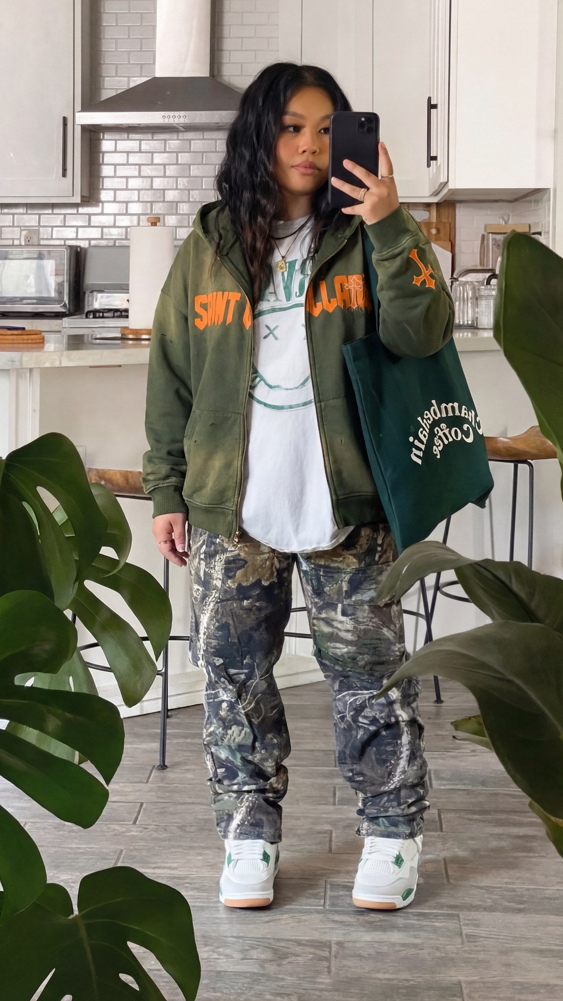 Woman wearing the oversized Saint Villains Redemption olive green zip hoodie open over a white tee with camo pants