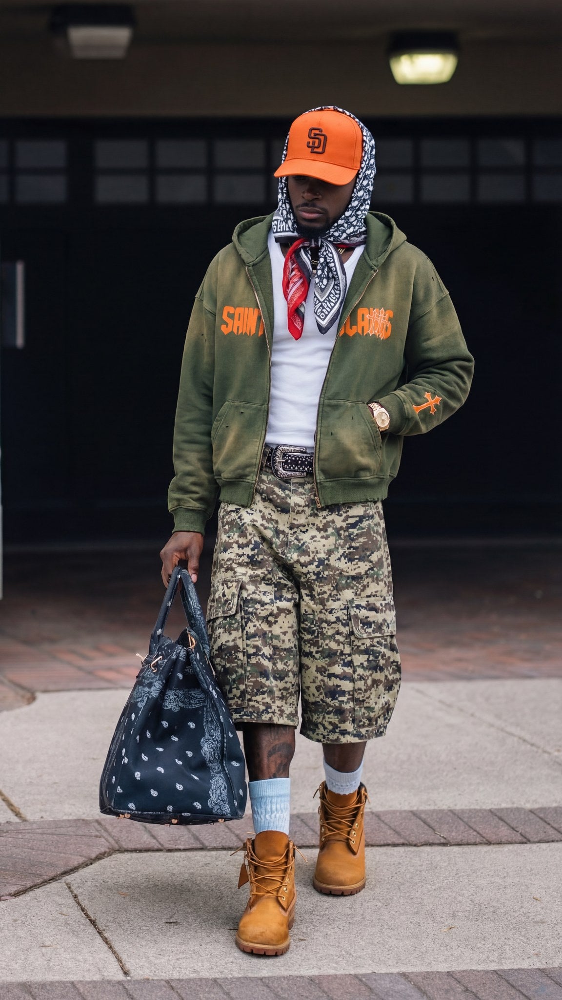 Man wearing the Saint Villains Redemption olive green zip hoodie with orange lettering, camo cargo shorts and Timberland boots