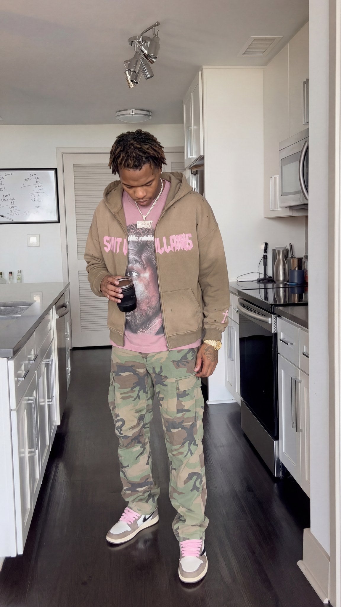 Man wearing the Saint Villains Redemption tan zip hoodie over a pink graphic tee with camo cargo pants